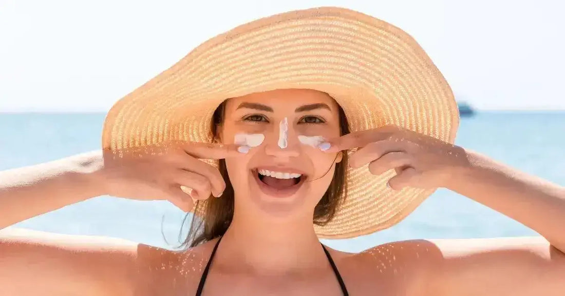 Smiling woman applying best Korean face sunscreens while wearing a sun hat at the beach.